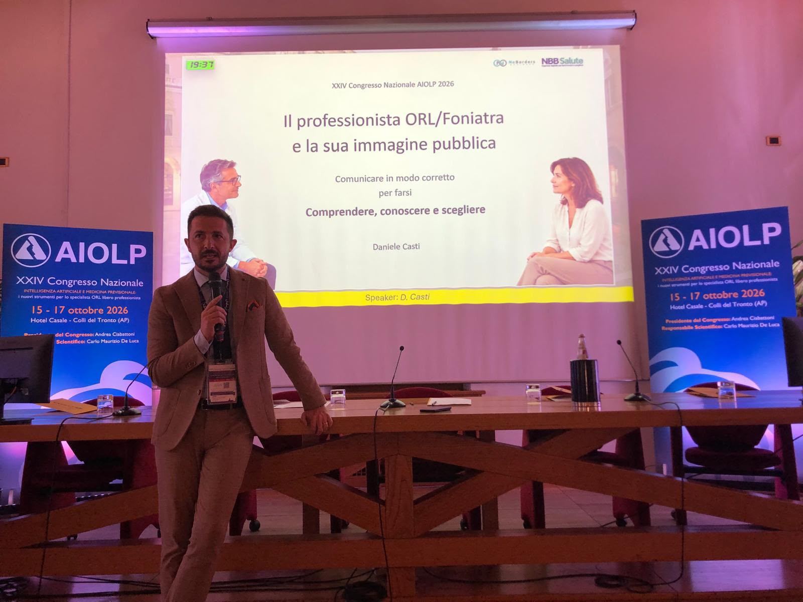 Daniele Casti con un microfono davanti allo schermo della presentazione e ai pannelli AIOLP.