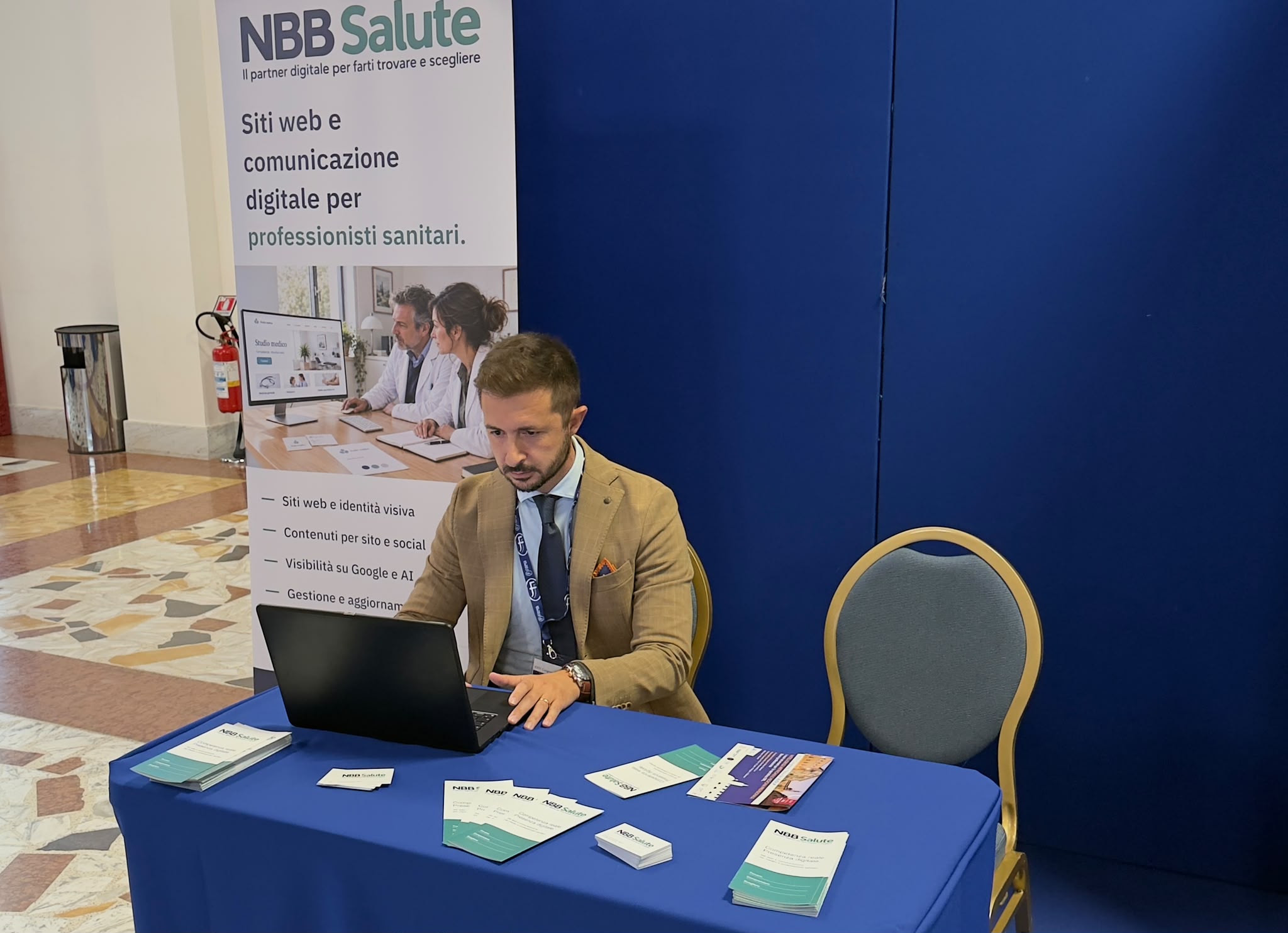 Postazione con un uomo seduto al computer, materiale informativo sul tavolo e un roll-up NBB Salute.